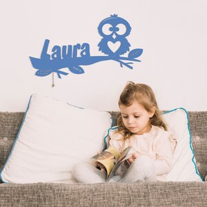 May include: A blue wooden owl with a heart on its chest is perched on a branch with the name "Laura" cut out of the wood. The owl and branch are mounted on a white wall. A young girl is sitting on a couch reading a book.