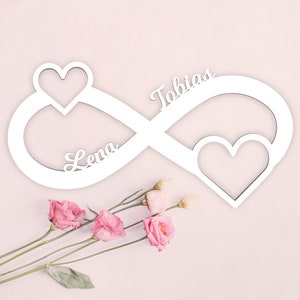 Peut inclure: Symbole d'infini en bois blanc avec les noms Lena et Tobias inscrits dessus. Deux cœurs sont découpés dans le symbole. Le symbole d'infini est sur un fond rose clair.