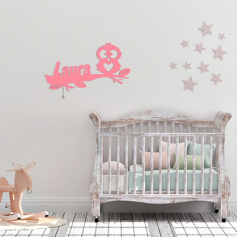 May include: Pink wooden owl with a heart and the name "Laura" on a branch. The owl is hanging on a white wall with pink stars. A white crib with a white and green blanket is in the background.
