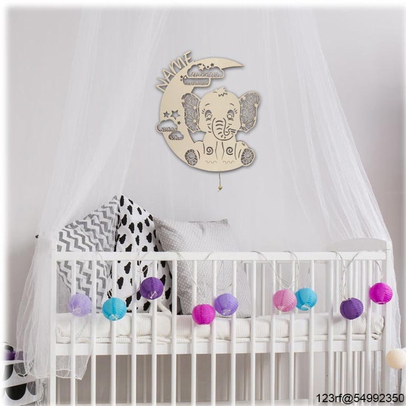 May include: A white wooden crescent moon with a cutout of an elephant and clouds. The moon has the word "NAME" written on it. The moon is hanging above a white cot with a white mattress and a white blanket. The cot is decorated with colourful paper lanterns.