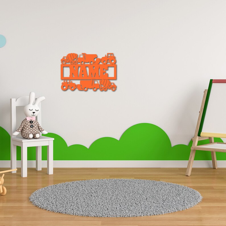 May include: Orange wooden wall decor with the word "NAME" surrounded by cutouts of construction vehicles. The decor is hanging on a white wall above a green and white rug.