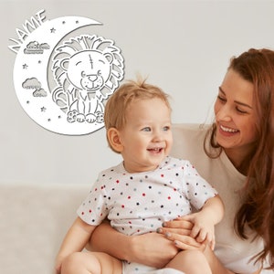 May include: A white wooden cutout of a crescent moon with stars and clouds, featuring a cartoon lion sitting on the moon. The cutout is hanging on a white wall behind a mother and child.