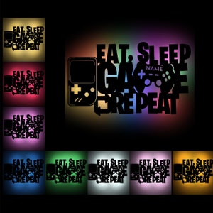 Könnte beinhalten: Ein schwarz-weißes Grafikdesign mit dem Text "EAT, SLEEP, GAME, REPEAT" mit einem Videospiel-Controller und einer Handheld-Spielkonsole. Die Grafik wird auf einem schwarzen Hintergrund mit einem Regenbogen-Verlaufseffekt angezeigt.