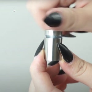 Puede incluir: Primer plano de un soporte de metal plateado con un inserto de vidrio transparente. El soporte est&aacute; siendo sostenido por una mano con esmalte de u&ntilde;as negro.