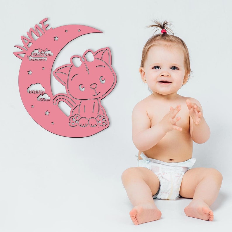 May include: Pink wooden cutout of a crescent moon with a cat sitting on it. The moon has stars and clouds with the word "Name" at the top. A baby girl is sitting in front of the cutout.