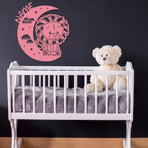 May include: A pink wooden lion and crescent moon growth chart with stars and clouds. The chart is mounted on a black wall above a white cot with a white teddy bear sitting inside.