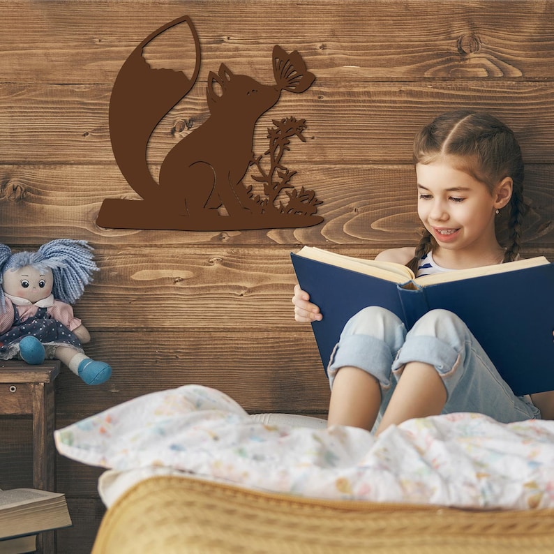 May include: A brown wooden wall decor featuring a fox silhouette with a butterfly, flowers, and leaves. A young girl is sitting on a bed reading a book.
