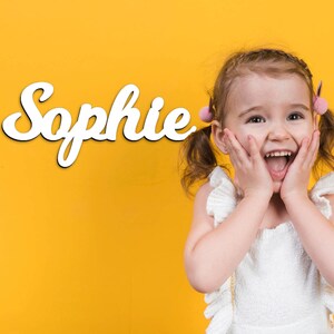 May include: A young girl with pigtails smiles excitedly in front of a yellow background with the name "Sophie" written in white cursive letters.