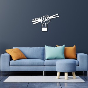 K&ouml;nnte beinhalten: Wei&szlig;e Wandkunst mit einer Faust, die Schlagst&ouml;cke h&auml;lt, auf denen das Wort "NAME" steht. Die Kunst wird an einer Wand &uuml;ber einem blauen Sofa mit orangefarbenen und blauen Kissen ausgestellt.