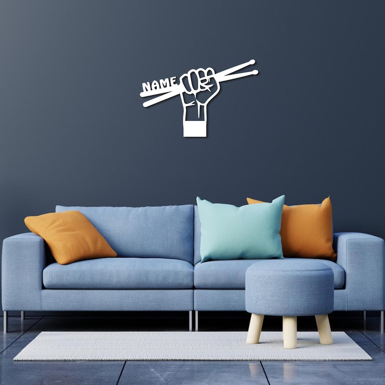 K&ouml;nnte beinhalten: Wei&szlig;e Wandkunst mit einer Faust, die Schlagst&ouml;cke h&auml;lt, auf denen das Wort "NAME" steht. Die Kunst wird an einer Wand &uuml;ber einem blauen Sofa mit orangefarbenen und blauen Kissen ausgestellt.