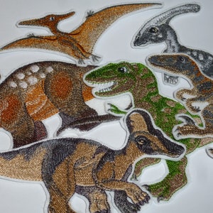Könnte beinhalten: Gestickte Dinosaurier-Patches in verschiedenen Farben, darunter Braun, Grün und Grau. Die Patches zeigen verschiedene Dinosaurierarten, wie einen Pterodactylus, einen Parasaurolophus und einen Tyrannosaurus Rex. Geeignet für Kleidung oder Accessoires.