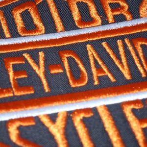 Könnte beinhalten: Nahaufnahme eines schwarzen Aufnähers mit orangefarbenem, gesticktem Text. Der Text lautet "MOTOR HARLEY-DAVIDSON CYCLES" in einer stilisierten Schriftart. Weiße Nähte umranden den orangefarbenen Text und begrenzen den Aufnäher.