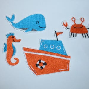 Könnte beinhalten: Eine Sammlung von aufgestickten Aufnähern mit Meeresbewohnern und einem Boot. Enthält einen blauen Wal, eine orangefarbene Krabbe, ein Seepferdchen und ein Boot mit Rettungsring. Die Aufnäher sind in Blau- und Orangetönen gehalten.