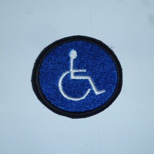 Könnte beinhalten: Gestickter Aufnäher mit dem internationalen Symbol für Barrierefreiheit. Das Design zeigt ein weißes Rollstuhlsymbol auf blauem, kreisförmigem Hintergrund, umrandet von einem schwarzen Rand. Dieser Aufnäher ist zum Aufnähen oder Aufbügeln.