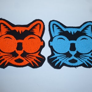Könnte beinhalten: Zwei aufgestickte Aufnäher mit einem Katzenkopf mit Brille. Einer ist orange auf schwarz, der andere blau auf schwarz. Die Katze trägt eine runde Brille und lächelt. Diese Aufnäher sind für Kleidung oder Accessoires.