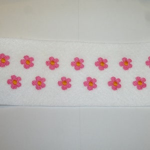 Könnte beinhalten: Ein weißes, rechteckiges Stoffstück mit gebogenem oberem Rand, bestickt mit einem sich wiederholenden Muster aus rosa Blumen mit gelben Zentren. Die Blumen sind gleichmäßig über dem Stoff verteilt.