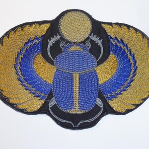 Könnte beinhalten: Ein gestickter Aufnäher mit einem blauen Skarabäus-Käfer mit goldenen und blauen Flügeln auf schwarzem Hintergrund. Die Flügel sind mit goldenen Perlen verziert, und der Körper des Käfers ist leuchtend blau. Über dem Käfer befindet sich eine goldene Sonne.