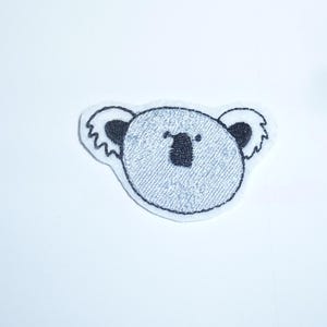Könnte beinhalten: Ein Koala-Bären-Aufnäher mit hellgrauem Körper und schwarzen Akzenten. Das Gesicht des Koalas ist mit einem strukturierten, denimartigen Stoff bestickt, und die Ränder sind weiß umrandet. Die Ohren sind schwarz mit gewelltem Rand.