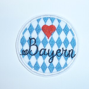 Könnte beinhalten: Runder, gestickter Aufnäher mit weißem Rand. Er zeigt ein blau-weißes Rautenmuster, ein rotes Herz und das Wort "Bayern" in schwarzer Schrift. Das Design befindet sich auf weißem Hintergrund.