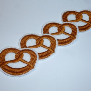 Könnte beinhalten: Vier gestickte Brezel-Patches. Jeder Patch zeigt ein detailliertes Brezel-Design in Brauntönen, umrandet von einem weißen Rand. Die Patches sind diagonal vor einem weißen Hintergrund angeordnet.