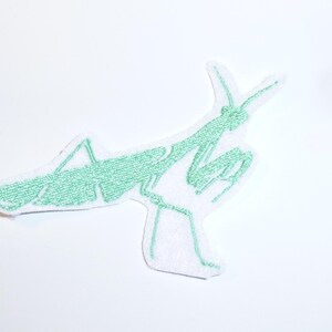 Könnte beinhalten: Hellgrüner, gestickter Aufnäher in Form einer Gottesanbeterin. Das Insekt ist im Profil dargestellt, mit seinen langen Beinen und Fühlern. Der Aufnäher hat einen weißen Hintergrund.