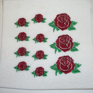 Könnte beinhalten: Quadratischer weißer Stoff mit gestickten roten Rosen und grünen Blättern. Die Rosen sind unterschiedlich groß, einige klein und andere größer. Die Stickerei ist detailliert und zeigt die Blütenblätter und Blätter. Der Stoff ist schlicht.