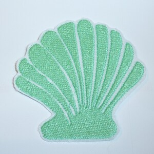 Könnte beinhalten: Ein hellgrüner Muschelförmiger Aufnäher mit weißem Rand. Der Aufnäher hat ein strukturiertes Aussehen und ein fächerartiges Design, wobei jeder Abschnitt der Schale nach außen strahlt. Der Hintergrund ist schlicht weiß.