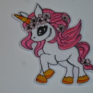 Könnte beinhalten: Gestickter Aufnäher eines weißen Einhorns mit rosa Haaren und Flügeln, einem gelb-orangen Horn und einem Blumenkranz. Das Einhorn befindet sich in einer Laufpose mit gelben Hufen. Der Aufnäher hat eine schwarze Umrandung.