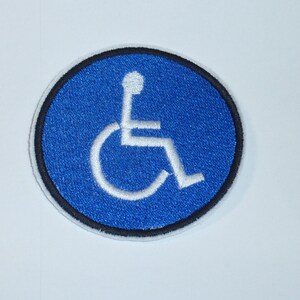 Könnte beinhalten: Runder, gestickter Aufnäher mit dem internationalen Symbol für Barrierefreiheit. Das Design zeigt einen weißen Rollstuhlfahrer in einem blauen Kreis, umrandet von Schwarz. Für Kleidung oder Accessoires.