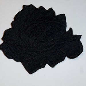 Könnte beinhalten: Ein schwarzer, bestickter Aufnäher in Form einer blühenden Rose. Das Design zeigt eine komplexe Fadenführung, die die geschichteten Blütenblätter der Rose bildet, mit einer dichten, strukturierten Oberfläche und einem definierten, gewellten Rand.