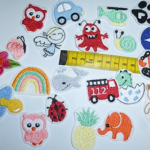 Könnte beinhalten: Eine Sammlung von aufgestickten Aufnähern in verschiedenen Formen und Farben, darunter eine Eule, ein Auto, ein Engel, ein Monster, eine Schnecke, ein Pfotenabdruck, ein Stern, ein Bagger, ein Dinosaurier, ein Bär, eine Blume, ein Regenbogen, ein Schnuller, ein Marienkäfer, eine Ananas und ein Elefant. Ein gelbes Maßband ist ebenfalls vorhanden.