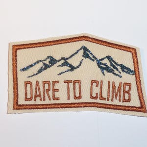 Könnte beinhalten: Rechteckiger Aufnäher mit einem Bergketten-Design und dem Schriftzug "DARE TO CLIMB". Der Aufnäher hat einen beigen Hintergrund, einen braunen Rand und die Berge sind blau.