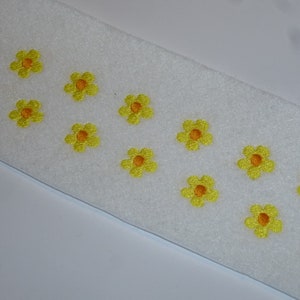 Könnte beinhalten: Ein weißes, rechteckiges Stoffstück, verziert mit gestickten gelben Blumen mit orangefarbenen Zentren. Die Blumen sind gleichmäßig über dem Stoff verteilt und bilden ein fröhliches und dekoratives Muster. Der Stoff scheint ein weiches, strukturiertes Material zu sein.