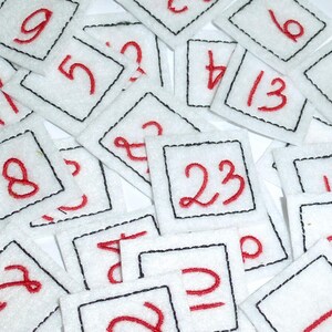 Könnte beinhalten: Eine Ansammlung weißer Filzquadrate mit rot gestickten Zahlen von 1 bis 23. Jedes Quadrat hat einen schwarz gestickten Rand. Die Quadrate sind verstreut, was ein spielerisches und lehrreiches Thema ergibt.