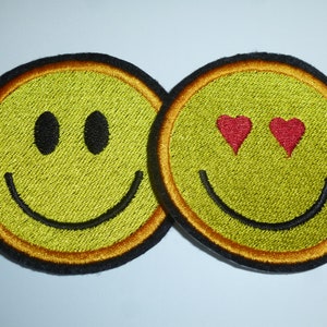 Könnte beinhalten: Zwei gestickte Aufnäher mit Smiley-Gesichtern. Einer hat schwarze ovale Augen, der andere rote Herzaugen. Beide sind gelb mit einem schwarzen Lächeln und einem schwarz-orangen Rand.