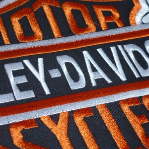 Könnte beinhalten: Gestickter Aufnäher mit dem Harley-Davidson-Logo in Orange, Weiß und Schwarz. Der schildförmige Aufnäher zeigt die Worte "MOTOR" und "CYCLES" in Orange und "HARLEY-DAVIDSON" in Weiß.