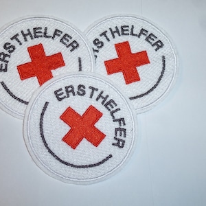 Könnte beinhalten: Drei weiße, kreisförmige Aufnäher mit einem roten Kreuz in der Mitte. Die Aufnäher haben schwarzen Text, der "ERSTHELFER" am Rand lautet. Die Aufnäher sind mit einer schwarzen Kontur bestickt.