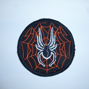 Könnte beinhalten: Runder schwarzer Aufnäher mit einer weißen Spinne und schwarzem Körper, vor einem orangefarbenen Spinnennetz-Design. Das Spinnennetz befindet sich auf einem schwarzen Hintergrund. Der Aufnäher hat einen schwarzen Rand.