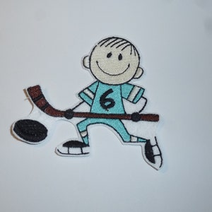 Könnte beinhalten: Gestickter Aufnäher mit einem Cartoon-Eishockeyspieler in einem türkisfarbenen Trikot mit der Nummer 6. Der Spieler lächelt und hält einen Hockeyschläger mit einem schwarzen Puck. Der Aufnäher ist weiß mit schwarzen und braunen Akzenten.