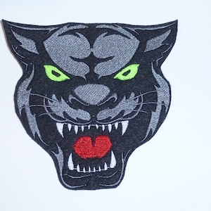 Könnte beinhalten: Gestickter Aufnäher mit einem grimmigen Pantherkopf. Der Aufnäher hat einen schwarzen Hintergrund mit grauen Details, grünen Augen und einem roten Maul. Die Zähne des Panthers sind weiß. Geeignet für Jacken, Taschen oder andere Stoffartikel.