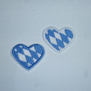 Könnte beinhalten: Zwei herzförmige Aufnäher mit blau-weißem Rautenmuster. Die Aufnäher haben einen blauen Rand und eine weiße Rückseite. Das Design erinnert an ein traditionelles bayerisches Motiv.