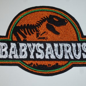 Könnte beinhalten: Gestickter Aufnäher mit einer Dinosaurier-Skelett-Silhouette in Schwarz auf orangefarbenem Hintergrund. Der Aufnäher hat einen schwarzen Rand mit grünen und weißen Akzenten und das Wort "BABYSAURUS" in Weiß.