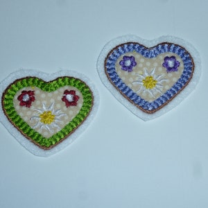 Könnte beinhalten: Zwei herzförmige, gestickte Aufnäher. Einer hat einen grünen Rand mit roten Blumen, der andere einen blauen Rand mit lila Blumen. Beide haben eine weiße Blume in der Mitte und einen beigefarbenen, gepunkteten Hintergrund.