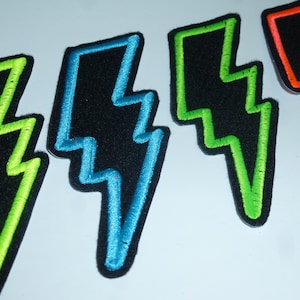 Könnte beinhalten: Nahaufnahme von vier gestickten Blitz-Patches. Jeder Patch hat eine schwarze Filzbasis mit einem kontrastierenden Neon-Umriss: grün, blau, grün und orange. Die Patches sind in einer Reihe angeordnet und zeigen die lebendigen Farben und das Design.