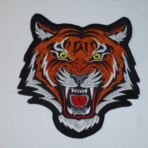 Könnte beinhalten: Gestickter Tigerkopf-Aufnäher mit orangefarbenen, schwarzen und weißen Details. Der Tiger hat gelbe Augen und einen geöffneten Mund, der Rot zeigt. Der Aufnäher ist schwarz umrandet.