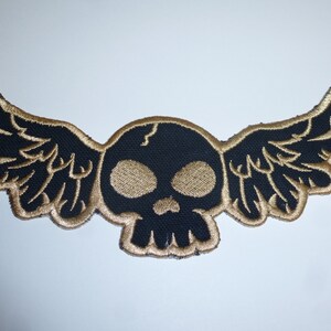 Könnte beinhalten: Gestickter Aufnäher mit einem schwarzen Totenkopf mit goldfarbenen Augen und einem Riss auf der Stirn, verziert mit goldfarbenen Flügeln. Der Aufnäher ist für das Aufnähen auf Kleidung oder Accessoires konzipiert.