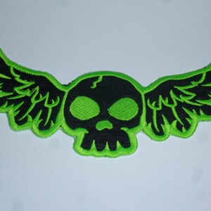 Könnte beinhalten: Gestickter Aufnäher mit einem schwarzen Totenkopf mit grünen Augen und einem Riss auf der Stirn, flankiert von schwarzen Flügeln. Das Design ist in leuchtendem Grün umrandet, was einen auffälligen Kontrast erzeugt. Ideal für Jacken, Taschen oder andere Stoffartikel.