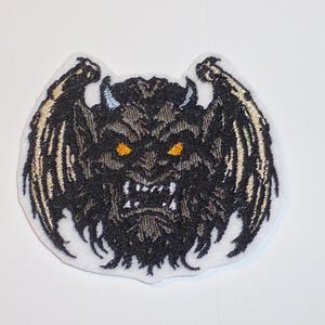 Könnte beinhalten: Gestickter Aufnäher mit einem stilisierten, wilden Gargoyle-Design. Der Aufnäher hat ein Schwarz- und Grauschema mit goldenen Akzenten auf den Flügeln und Augen. Der Aufnäher befindet sich auf weißem Hintergrund.