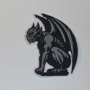 Könnte beinhalten: Gestickter Aufnäher mit einem schwarzen und grauen Wasserspeier in sitzender Position mit großen Flügeln. Der Aufnäher hat einen weißen Rand und ist zum Aufbügeln oder Aufnähen auf Stoff vorgesehen. Der Wasserspeier ist ein gotisches Architekturelement.