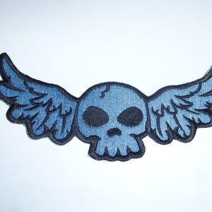 Könnte beinhalten: Blauer Totenkopf-Aufnäher mit Flügeln aus Jeansstoff. Der Totenkopf hat schwarze Augenhöhlen und eine schwarze Umrandung. Die Flügel sind weit ausgebreitet, auf weißem Hintergrund.